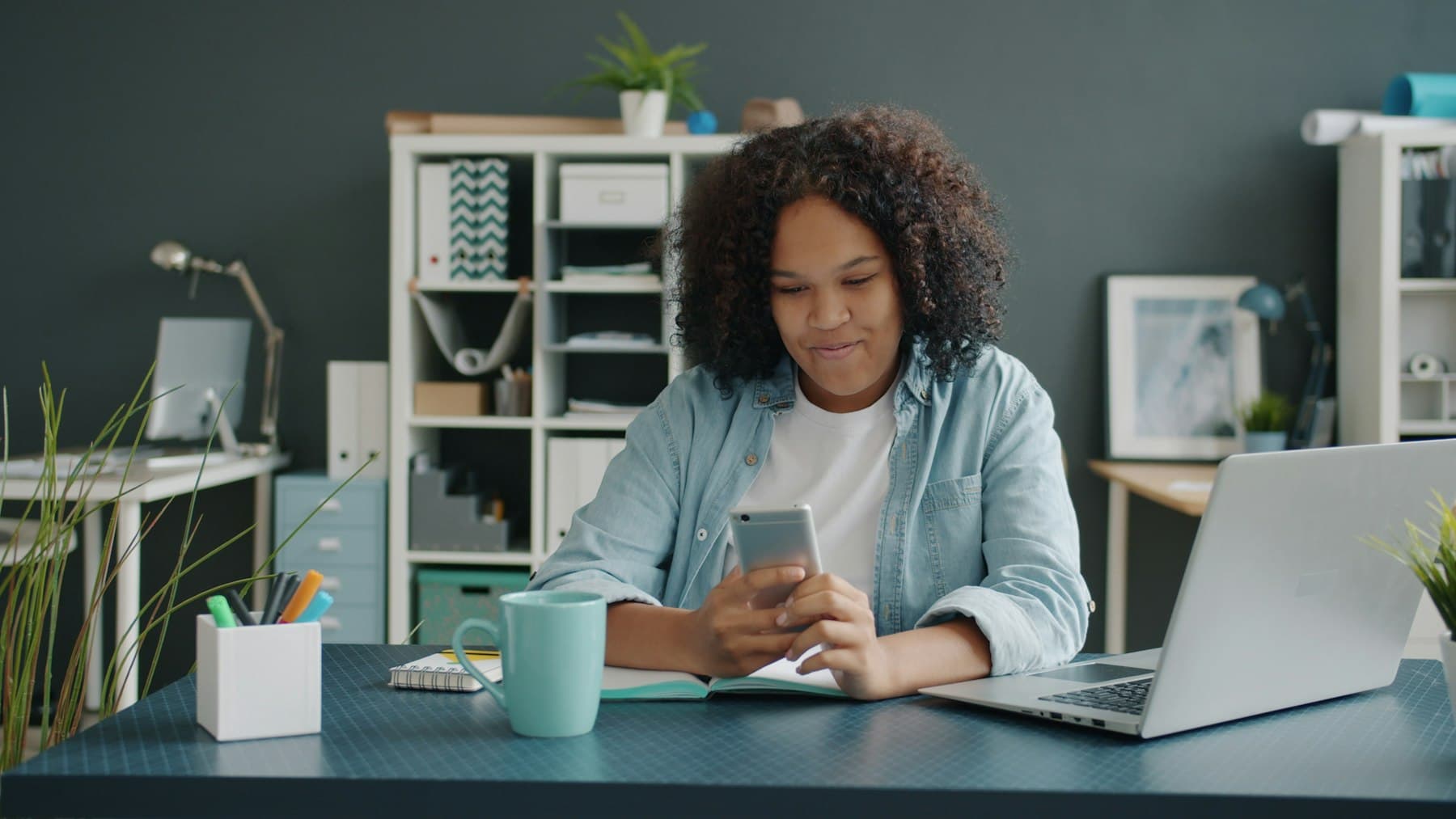 A woman checking her phone at her desk, in front of her laptop